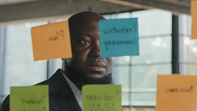 African american analyst serious businessman project manager check schedule read information on reminders stickers look at glassy wall with colorful sticky notes managing goals daily corporate tasks