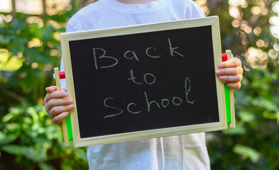 child boy in a white shirt, holds in his hands a small black chalkboard in front of him with the inscription back to school
