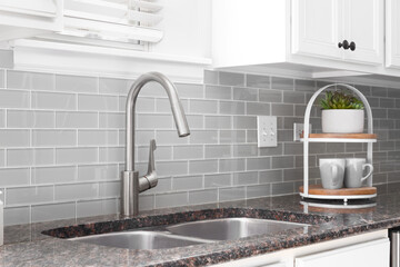 A kitchen sink detail shot with white cabinets, grey subway tile backsplash, and a granite...