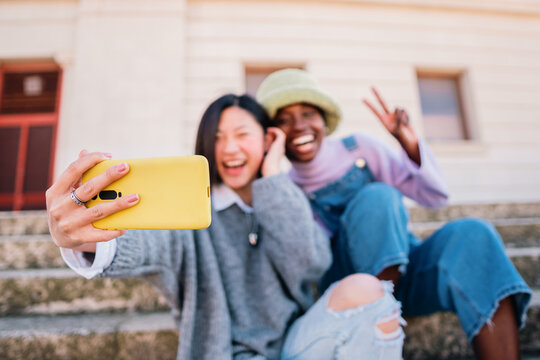 Two joyful cheerful girls taking a selfie while sitting together outdoors and showing peace gesture