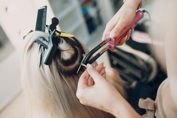 Hairdresser female making hair extensions to young woman with blonde hair in beauty salon....