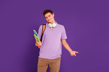 Young happy man student in casual clothes posing with backpack and notebooks, isolated on studio violet background. Education concept.