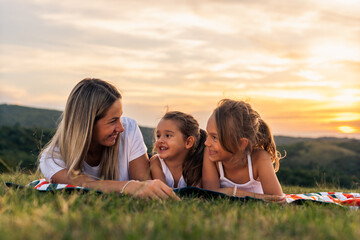 Happy mother and daughters smiling and laughing lying on blanket at the lawn in summer park. Family spend their free time together on vacation.