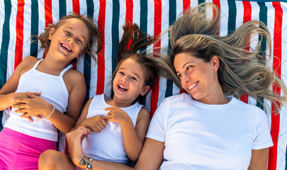 Happy mother and daughters smiling and laughing lying on blanket at the lawn in summer park. Family spend their free time together on vacation.