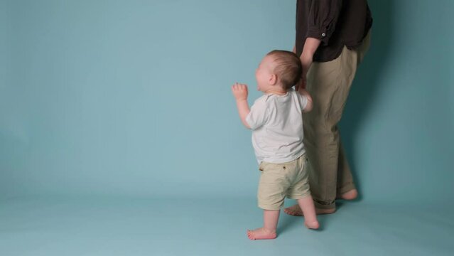 Happy toddler baby learns to walk and takes his first steps with the help mother. Child boy walks with his mom for the first time on a studio blue background. Kid age one year, slow motion