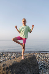 The girl does yoga on the beach, keeps balance standing on 1 leg.