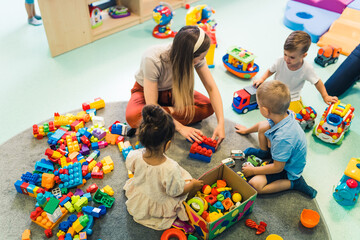 Playtime at nursery school. Toddlers with their teacher sitting on the floor and playing with building blocks, colorful cars and other toys. High quality photo - obrazy, fototapety, plakaty