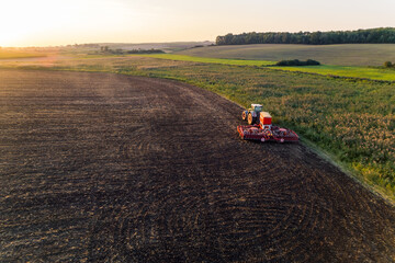 Tractor with sowing machine driving through brown field with greenery on the side and in the fading background. Morning landscape. Horizontal shot. High quality photo