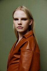 Handsome pretty blonde stylish young lady in brown palette leather dress serious looking over her shoulder at camera posing isolated on over olive green studio wall background. Fashion portrait banner