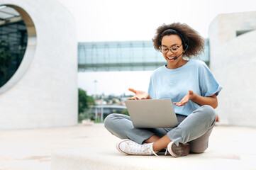 Positive friendly african american curly young woman with headset, manager, coach, having online consultation, negotiations with colleagues, sits outdoors, using laptop, talking on video call, smiling