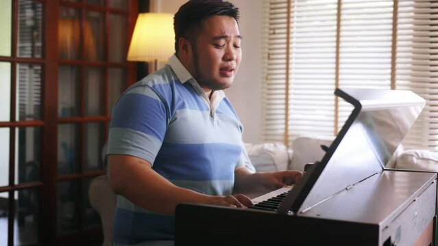 Young Asian chubby man singing and playing piano. Plus-size man practicing electric keyboard instrument at home. High quality 4k footage