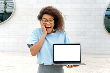 Excited shocked amazed african american young woman with curly hair, stylishly dressed, stands outdoors, holds an open laptop with blank white mock-up screen in hand, looks surprised at the camera