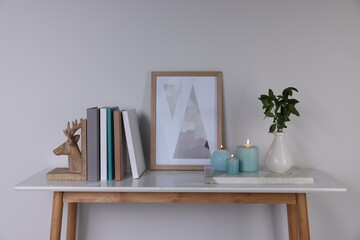 Burning candles, books, picture and vase with green branches on console table near white wall
