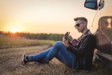 Naklejka na ściany i meble Musician with guitar sits on the dusty countryside road near the car in the sundown rays.