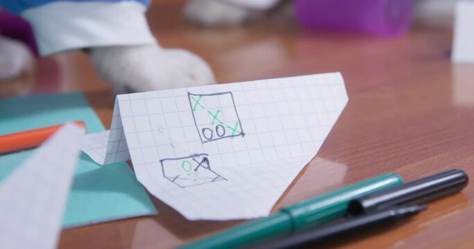 Paper airplane with a picture of a tic-tac-toe game is lying on a table, that someone is knocking on and therefore it is shaking, close up. Dog in the t-shirt put its paw on table