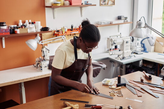 Portrait of black young woman cutting leather and creating handmade belt in workshop, copy space - Powered by Adobe