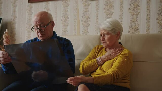 Concerned old caucasian grey-haired married couple holding x-ray results, scan of a man's lungs, and worrying about health while sitting on a beige leather sofa at home together. High quality 4k