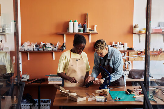 Portrait of black young woman learning leatherworking craft from artisan in workshop, copy space