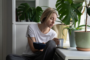 Upset single middle-aged female thinks about uneasy life taking mug with hot drink sitting near window. Blonde woman holds breakfast bowl searching job via smartphone put on windowsill closeup