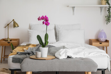 Tray with orchid flower and candle on bench near bed in room interior