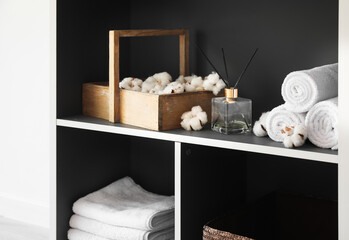 Shelving unit with towels, cotton flowers and reed diffuser near light wall