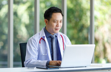 Asian successful professional middle aged male doctor in white lab coat with stethoscope sitting using laptop notebook computer working researching online in glass windows hospital clinical office