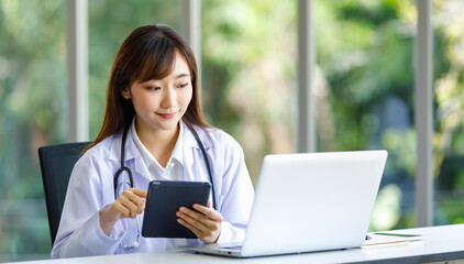 Asian successful professional young female doctor in white lab coat uniform with stethoscope sitting smiling using typing laptop notebook computer working researching online in hospital clinic office