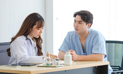 Asian young professional female doctor in white lab coat with stethoscope sitting writing down prescription on work desk while consulting with male patient in blue hospital uniform in clinic office