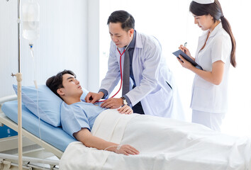 Asian professional male doctor in white lab coat using stethoscope listening monitoring heartbeat pulse on patient chest while female nurse in uniform writing symptom data on clipboard in ward room