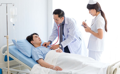 Obraz premium Asian professional male doctor in white lab coat using stethoscope listening monitoring heartbeat pulse on patient chest while female nurse in uniform writing symptom data on clipboard in ward room