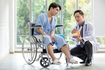 Asian middle aged professional male doctor in white lab coat with stethoscope sitting holding human foot anatomy model showing explaining to patient in blue uniform on wheelchair in hospital walkway
