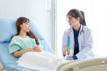 Fototapeta premium Asian young professional female doctor in white lab coat with stethoscope sitting holding human spine skeleton model showing explaining supporting comforting patient laying down on bed in ward room