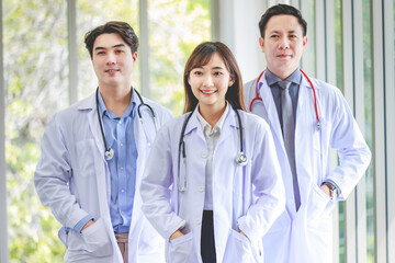 Closeup shot of Asian young cheerful happy professional intern practitioner doctor in white lab coat with stethoscope standing smiling in front of male and female colleagues on blurred background