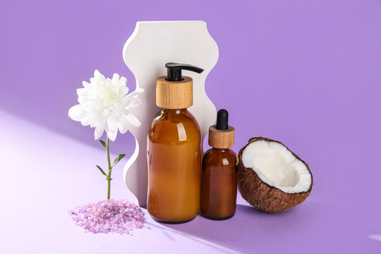 Composition with bottles of natural cosmetic products, coconut and chrysanthemum flower on lilac background