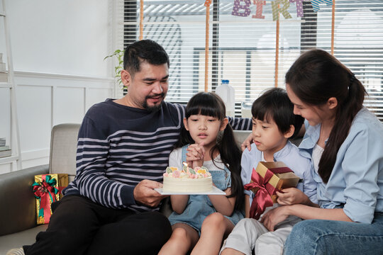 Happy Asian Thai family, young daughter is surprised with birthday cake and gift, blows candle, pray and cheerful celebrates party with parents together in living room, domestic home event lifestyle.