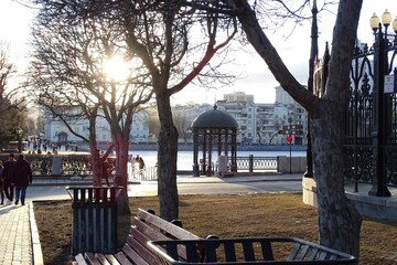 Horizontal full-color photo. Urban landscape. In the foreground is a vintage porch with cast iron elements. In the background - trees, buildings, lanterns, cars, sunset sky.