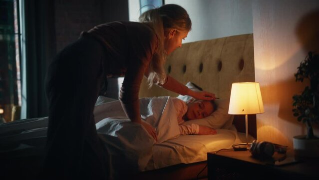 Mother Caring For Her Lovely Daughter, Putting Her To Sleep In Bed Before At Home In The Evening. Babysitter Tucking Little Girl In Blanket And Turning Off Light In Table Lamp.