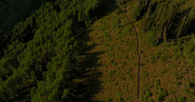 Aerial over backcountry mountain range as two bikers bike down trail at sunset.