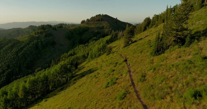 Aerial over backcountry mountain range as two bikers bike down trail at sunset.