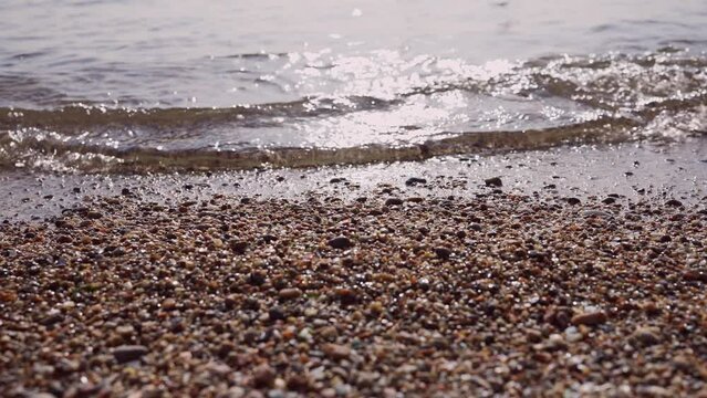 Close-up of pebble beach with transparent waves with sun glare on the water. Close up of stony sea shore. Natual background of calm sea in the surf zone. Camera moves sideway to the right side