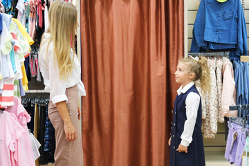 Mother and daughter are standing in the store opposite the fitting room. Girl trying on a school dress