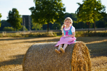 Cute little kid girl in traditional Bavarian costume in wheat field. Happy child with hay bale...