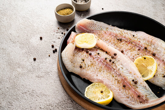 Raw pangasius fish fillet with lemon and spice in frying pan on concrete background