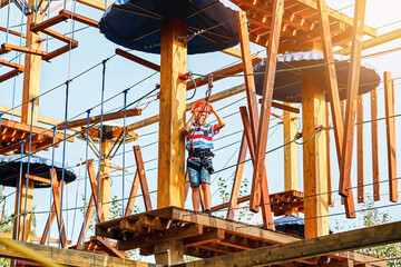 Boy in a helmet and climbing equipment standing on a wooden platform against the sky, spending time in rope park in summer evening