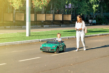 Mom controls children car with remote at walk with toddler child in public park. Toddler boy sits...