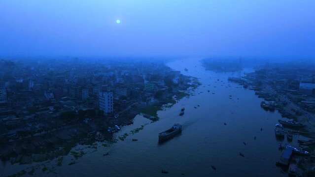 Aerial view of a riverside city in a full moonlit night. Cargo ship passing through the river in a misty gloomy weather. Plane pilot pov view.