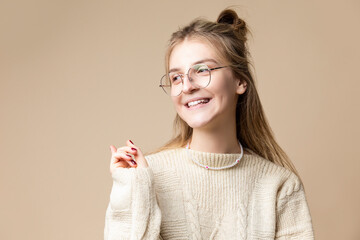 Smiling Caucasian Blond Female Wearing Knitter Decorated Warm Sweater And Glasses while Looking Aside with Lifted Finger Over Beige.