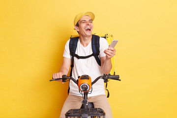 Indoor shot of happy young delivery guy holding cell phone, wearing cap and thermo bag, riding...