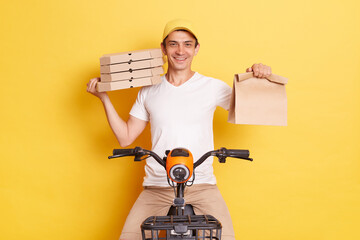 Indoor shot of delivery man riding bike and holding paper packages with takeaway food, wearing...
