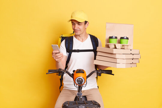 Indoor shot of young adult Caucasian delivery man using smartphone, checking address for delivering order, wearing cap and thermo bag, riding bicycle, isolated on yellow background